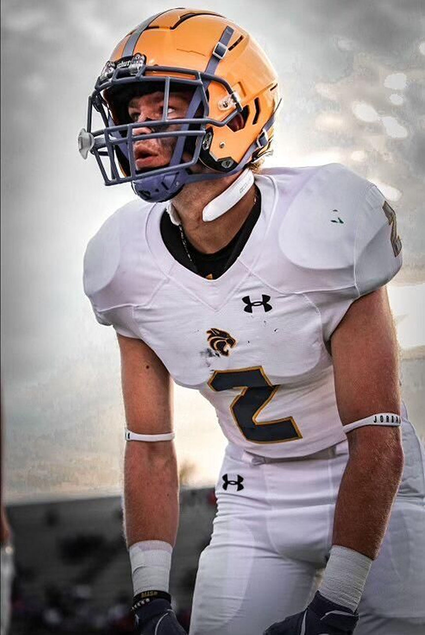 An American football player wearing a white jersey with the number 2 and a yellow helmet with a grey facemask. The player is in a ready stance on a football field, looking off to the side, wearing a white Q-Collar device around his neck.